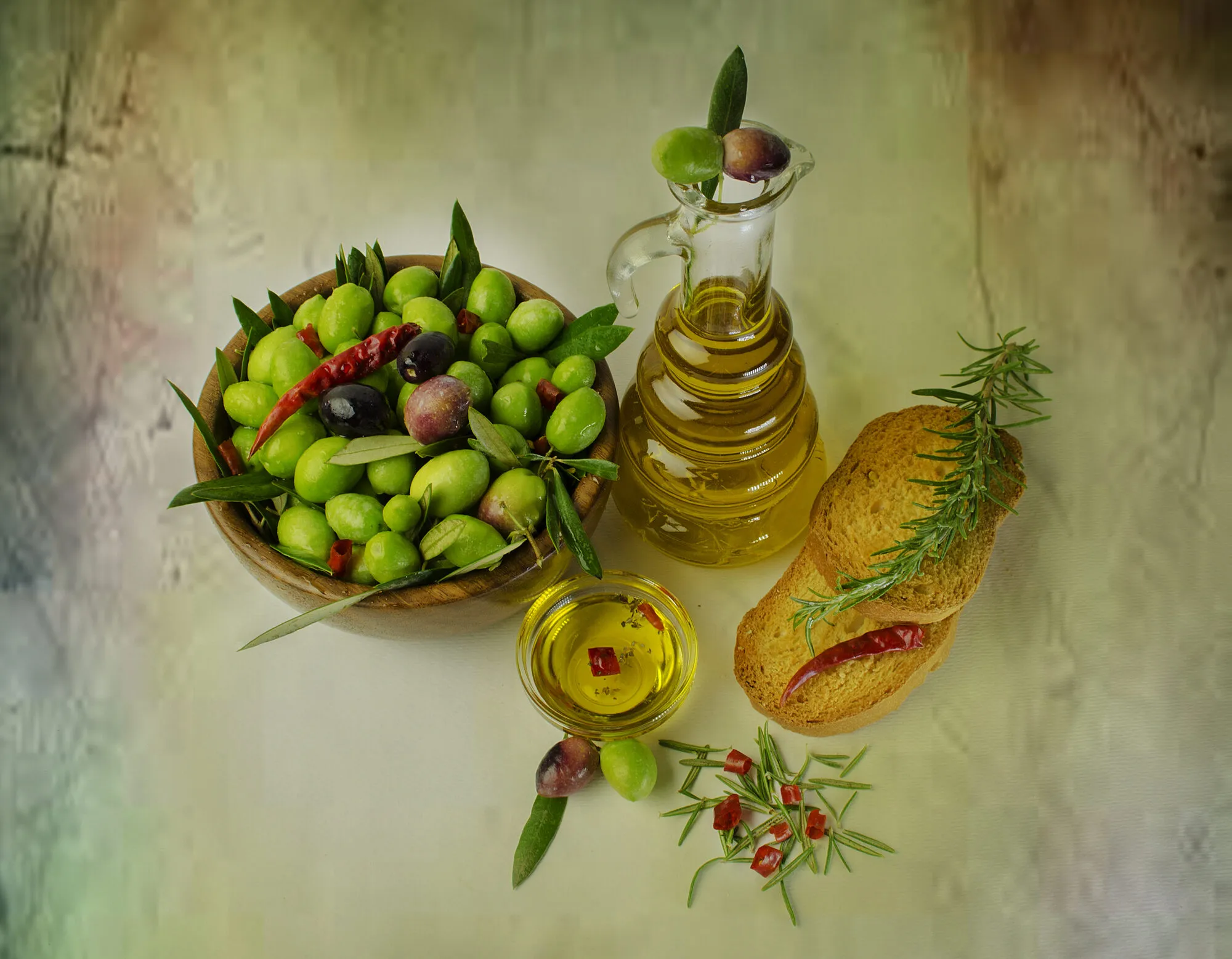 A wooden bowl of green olives beside a glass cruet of extra virgin olive oil and bread with rosemary